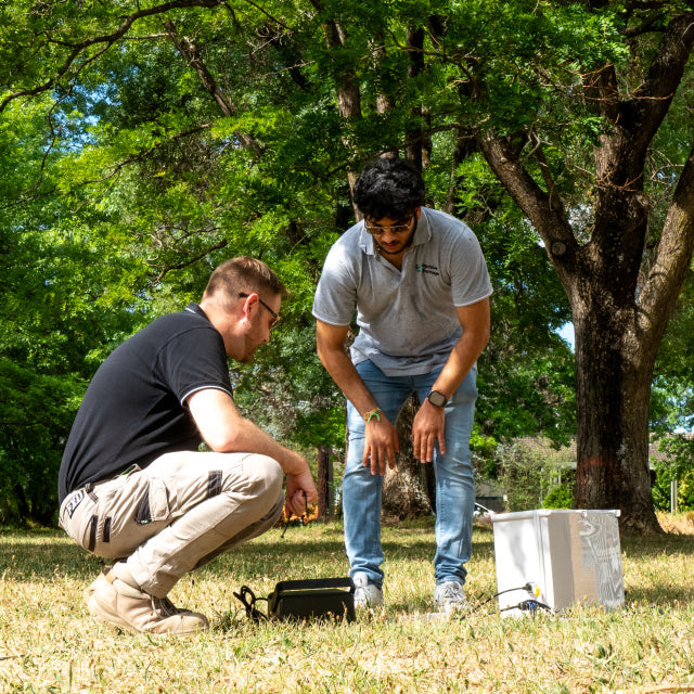 Two men working on a device in a park setting with trees and grass.