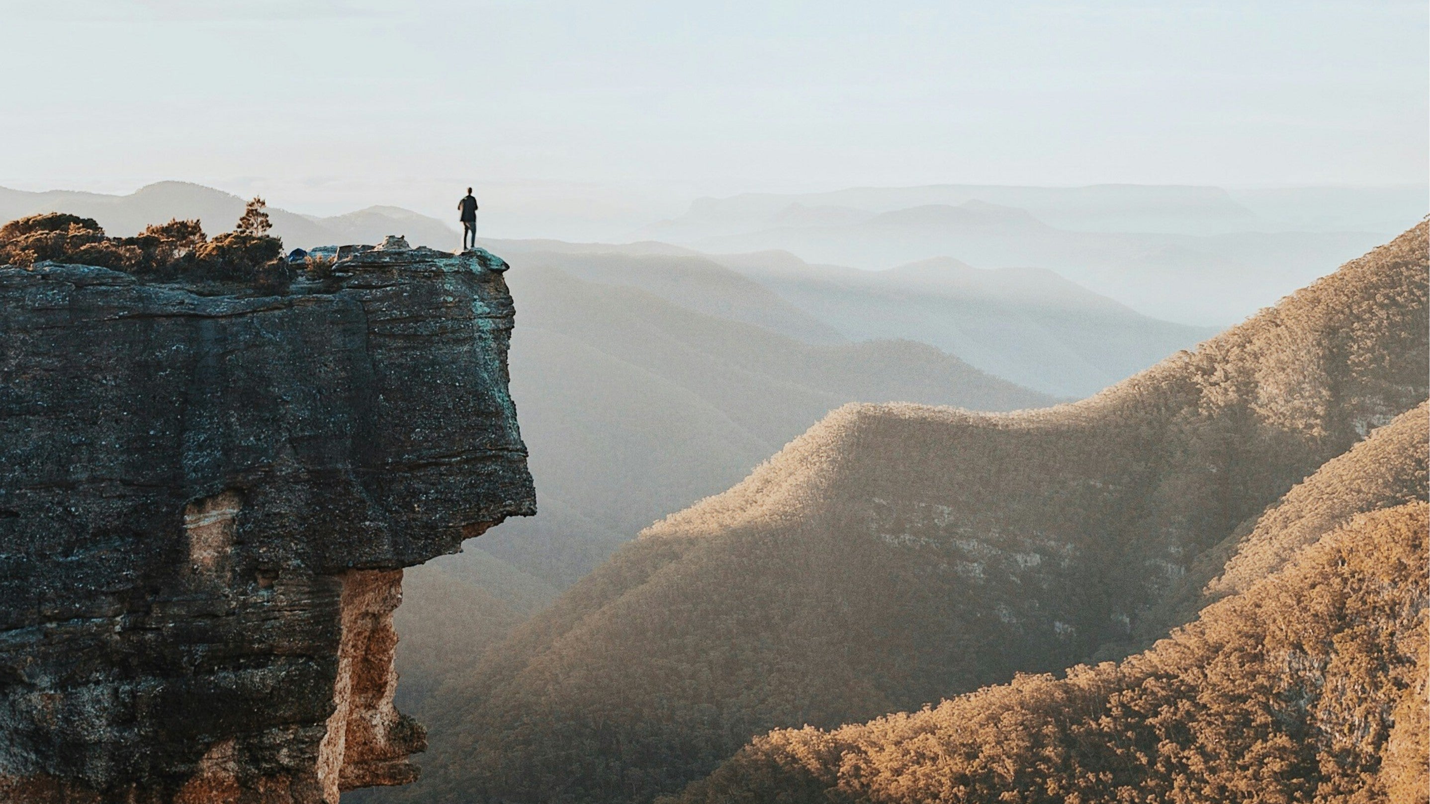 Person standing on a cliff overlooking a vast landscape with mountains and valleys.